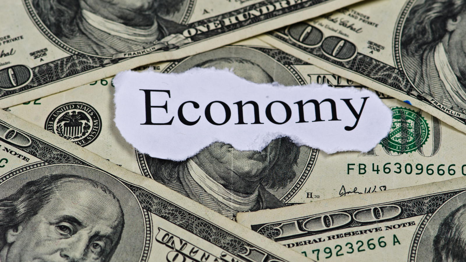Close up of 100 dollar bills in a pile and a torn out piece of paper laying on top of them with the printed word Economy.