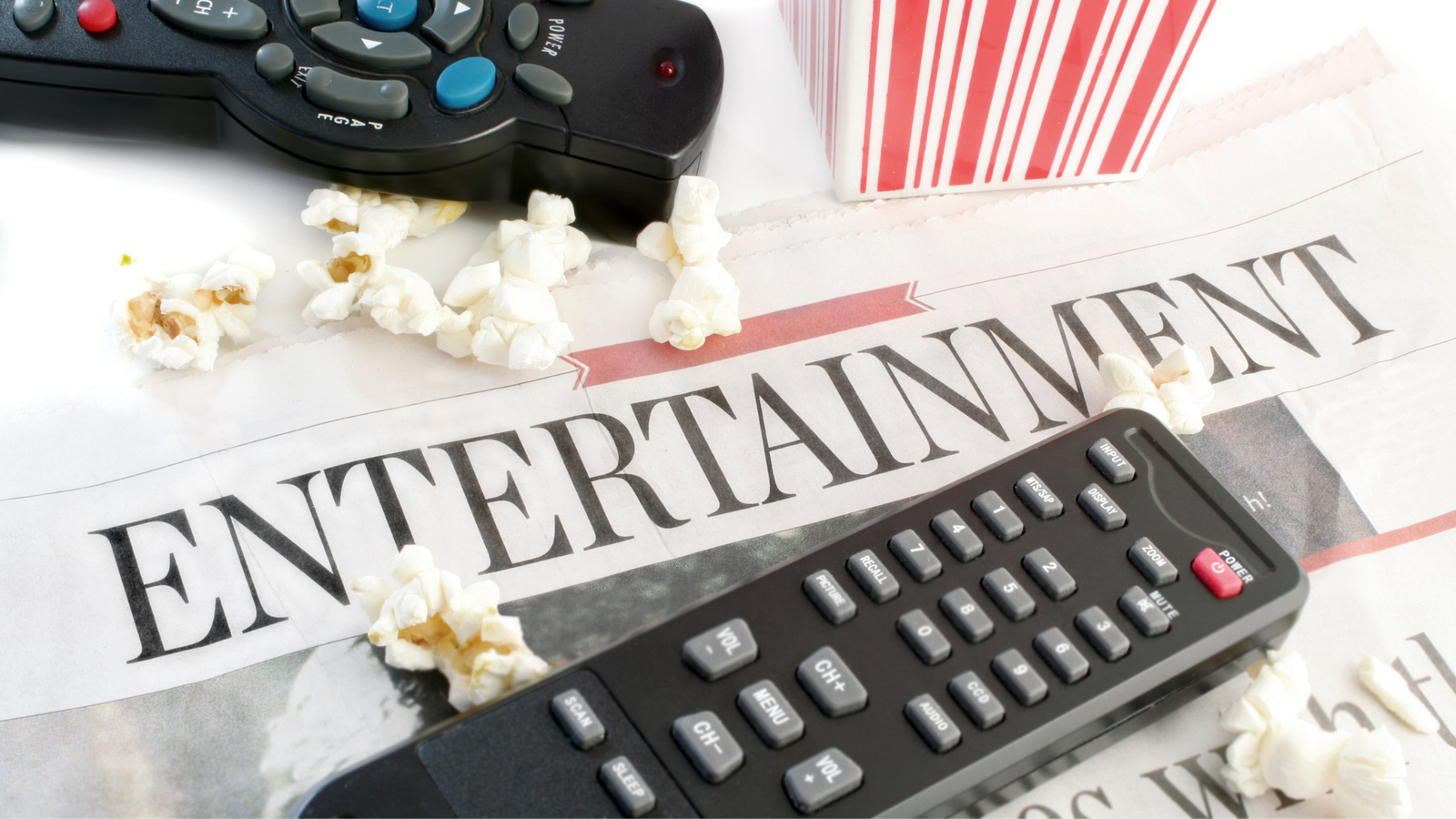 Close up of a newspaper with the large word ENTERTAINMENT across the top laying on a table with two remote controllers and popcorn.