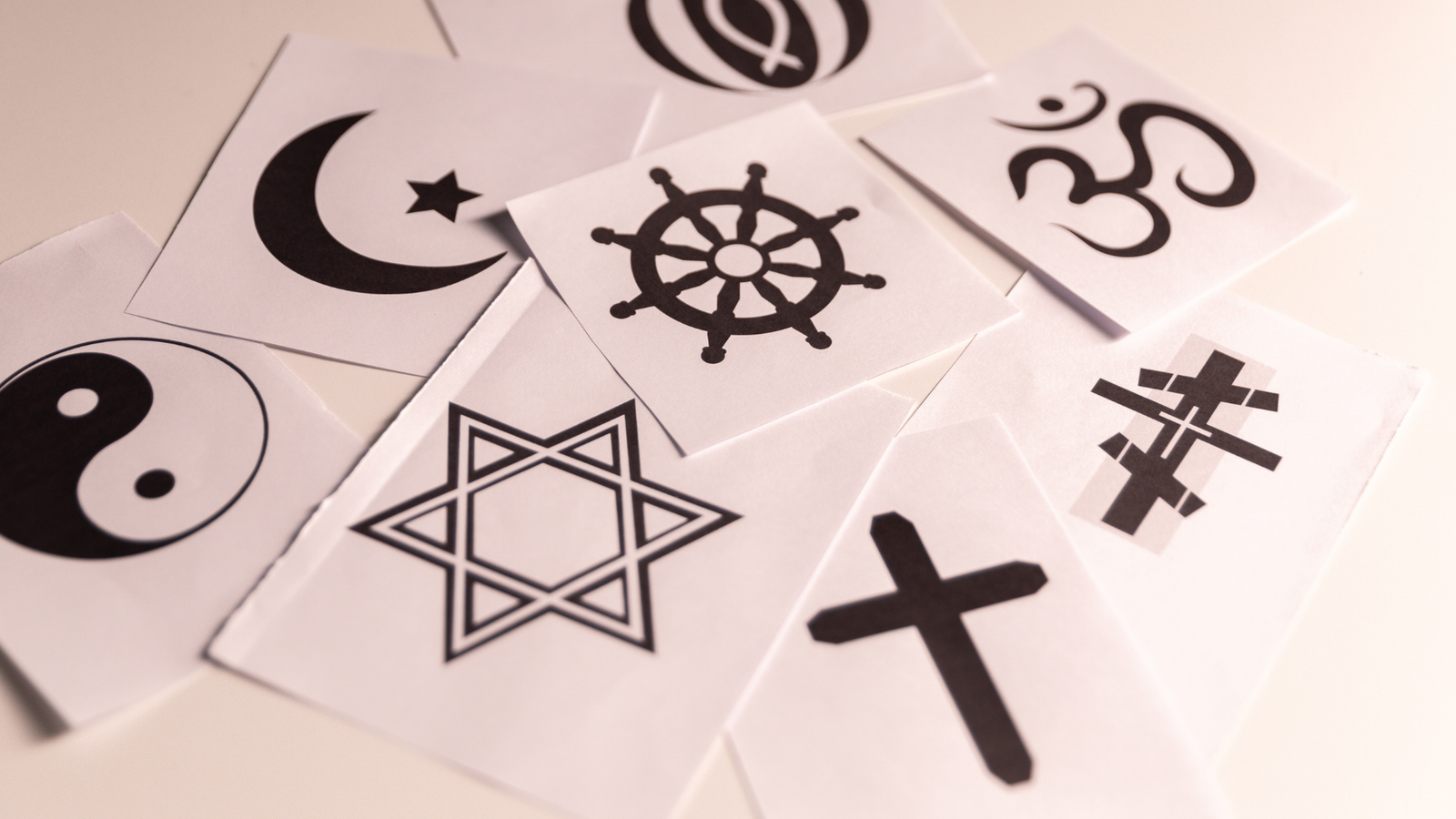 A collection of small white square papers scattered on a surface, each featuring a black religious or philosophical symbol, including a Star of David, a Crescent and Star, a Christian Cross, an Om symbol, a Dharma Wheel, and a Yin Yang.